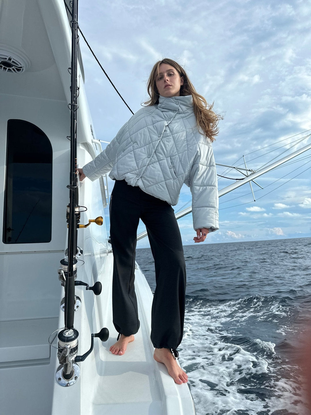 Model standing on a boat, wearing a light blue puffer jacket with a high collar and black pants, with the ocean and sky in the background.