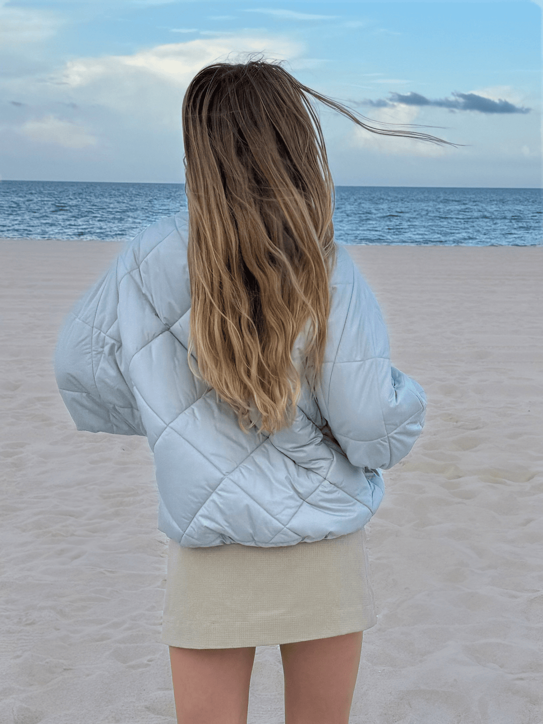 Back view of a model with long hair wearing a light blue puffer jacket and a beige skirt, standing on a sandy beach with the ocean and sky in the background.
