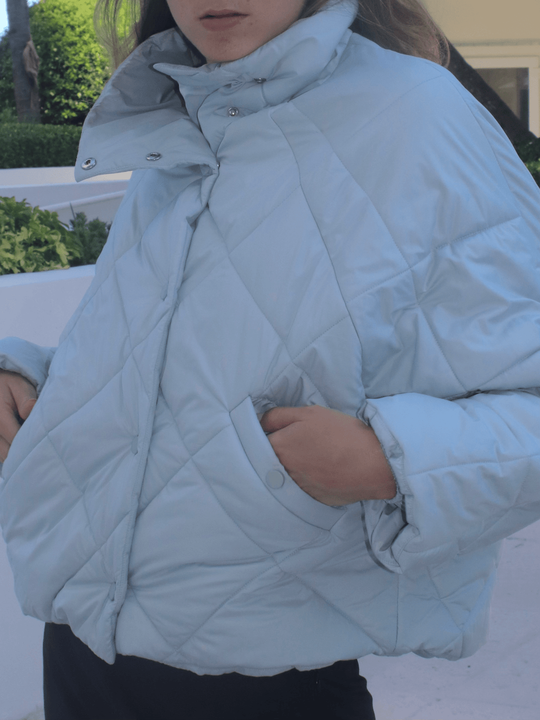 Front view of a model wearing a light blue puffer jacket with her hands in pockets, featuring a high collar and buttons, with greenery and white walls in the background.