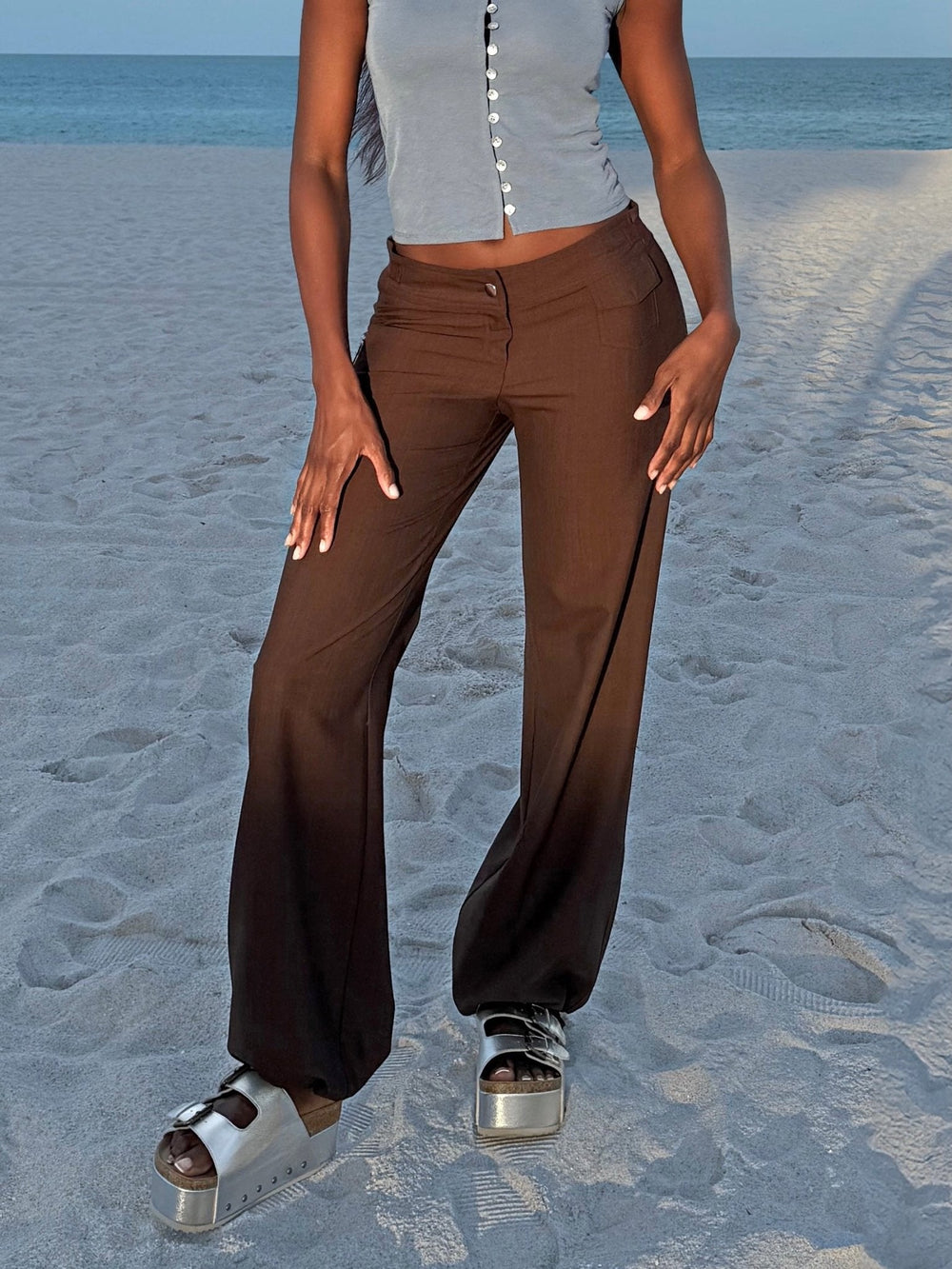 Model wearing brown wide-leg pants and platform sandals on a sandy beach, paired with a blue button-up top.