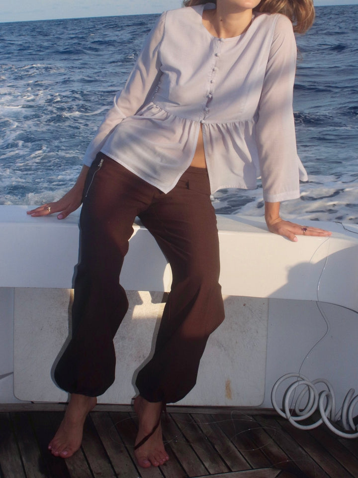 Model sitting on the edge of a boat deck wearing a light, flowy blouse with button details, paired with high-waisted brown relaxed-fit pants featuring drawstring-adjustable ankle cuffs. 