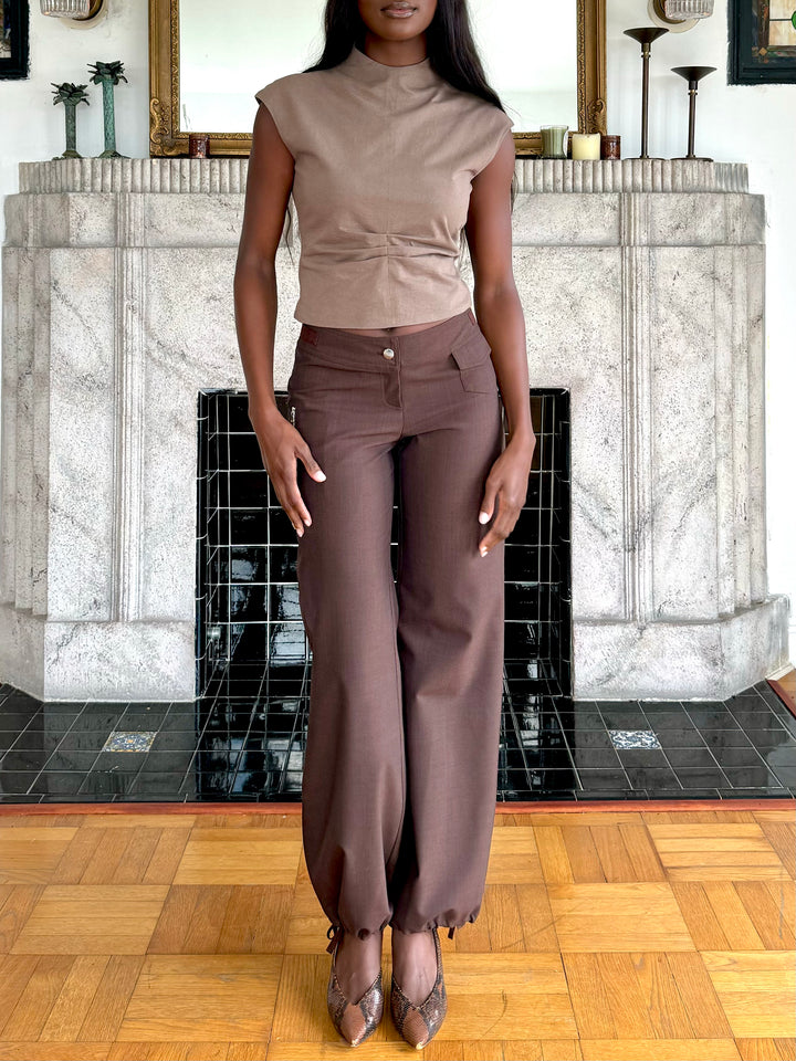 Model standing indoors wearing a sleeveless brown top and matching brown wide-leg pants with snakeskin-patterned heels, posing in front of a decorative fireplace.