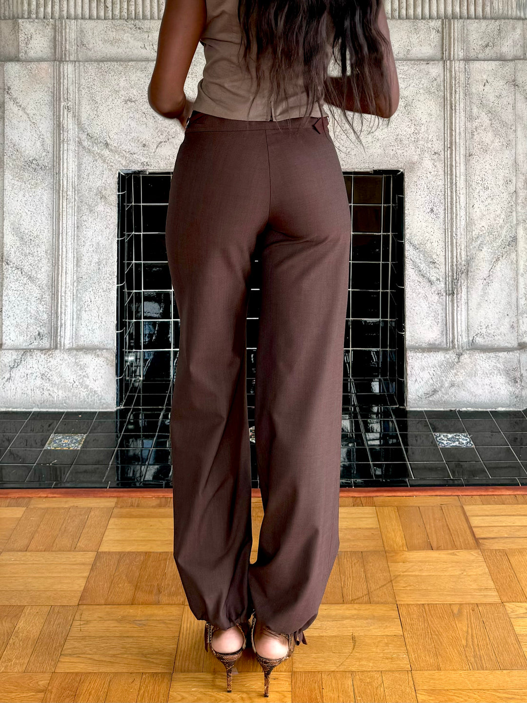 Back view of model wearing a  brown top and matching brown wide-leg pants with snakeskin-patterned heels, posing in front of a decorative fireplace.