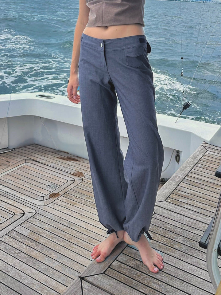 Model standing barefoot on a boat deck wearing high-waisted blue relaxed-fit pants with a button closure, drawstring-adjustable ankle cuffs, and a utility pocket detail. Paired with a cropped beige top.