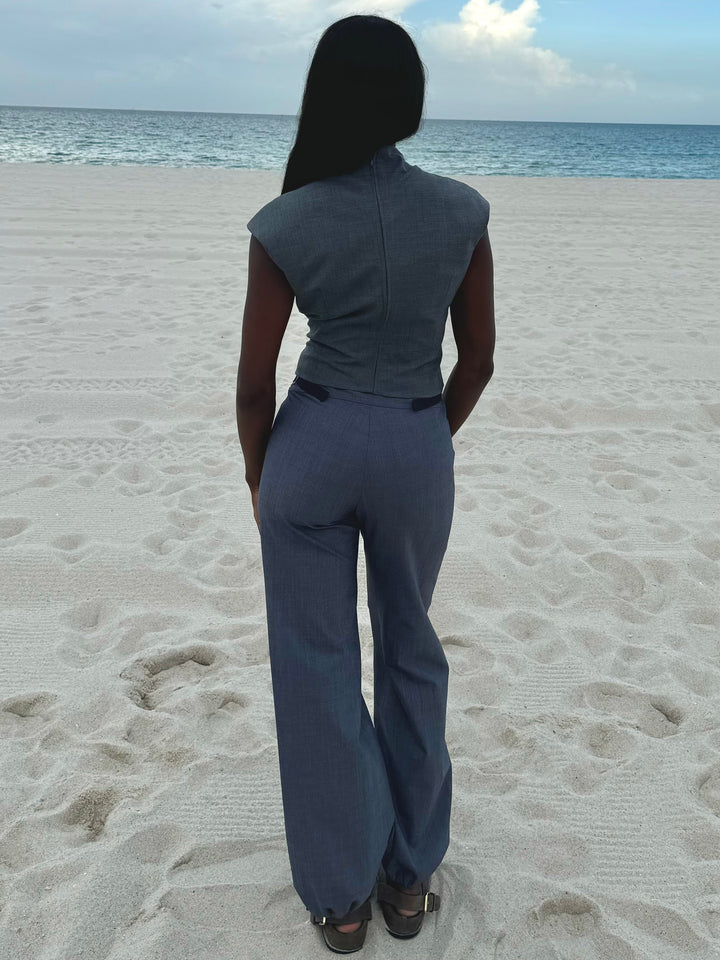 Back view of a model standing on a sandy beach, wearing a fitted gray sleeveless top and high-waisted blue relaxed-fit pants with drawstring-adjustable ankle cuffs.