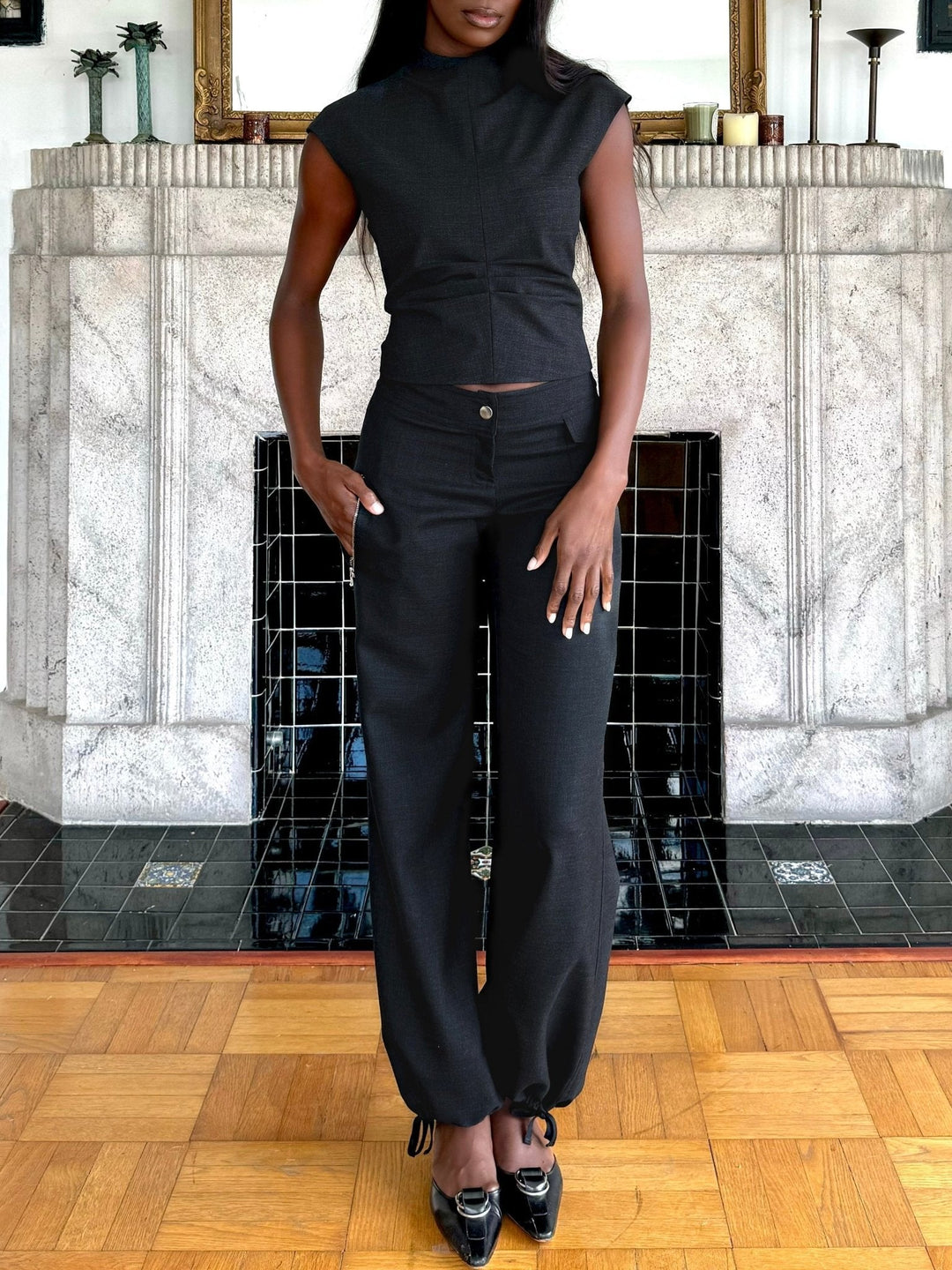 Model wearing a fitted black sleeveless top paired with black relaxed-fit pants that feature a button closure and drawstring-adjustable ankle cuffs. Styled with black pointed-toe heels, this outfit is both elegant and modern, set against a chic interior backdrop with a tiled fireplace.