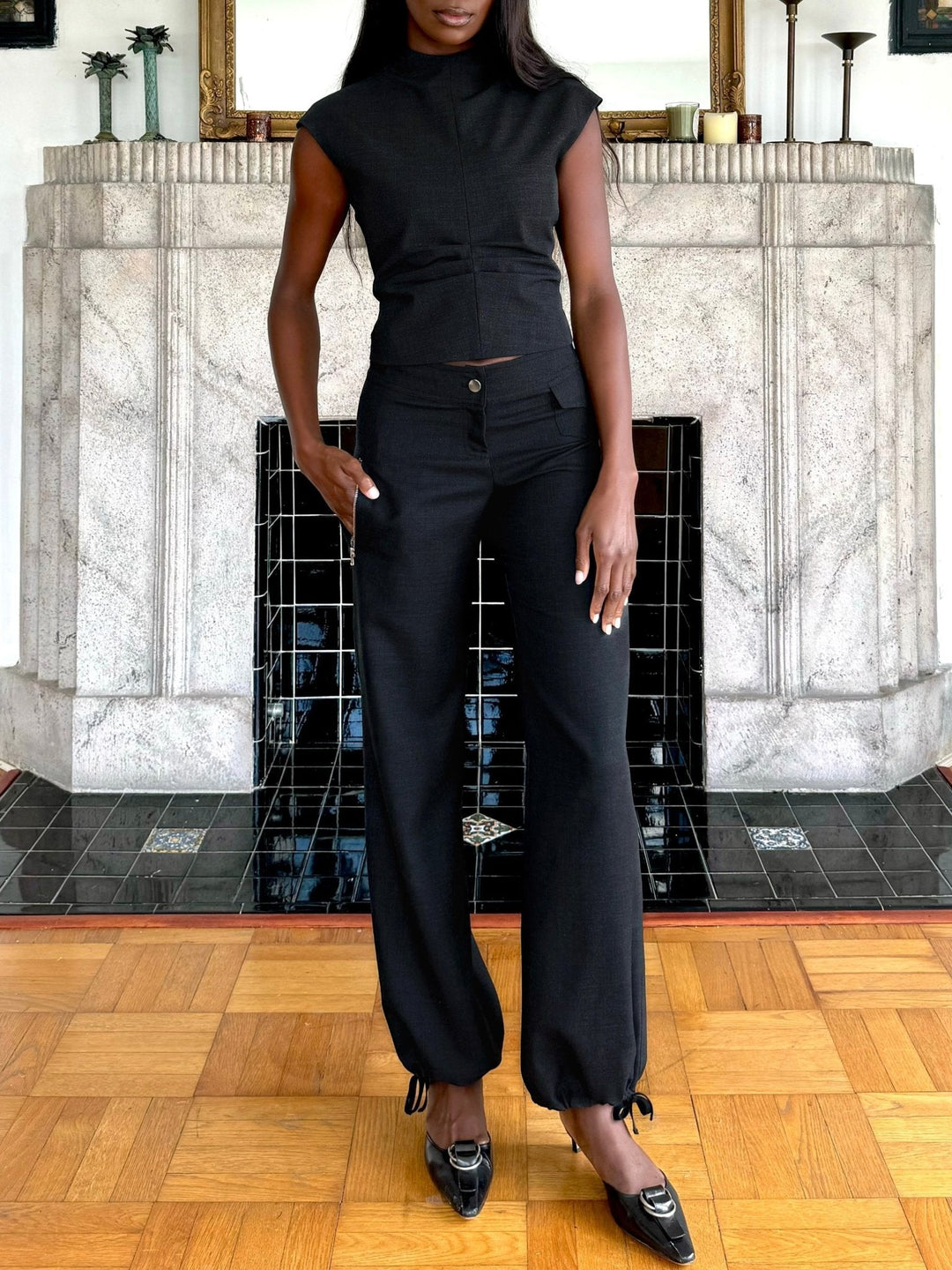 Model wearing a fitted black sleeveless top paired with black relaxed-fit pants that feature a button closure and drawstring-adjustable ankle cuffs. Styled with black pointed-toe heels, this outfit is both elegant and modern, set against a chic interior backdrop with a tiled fireplace.