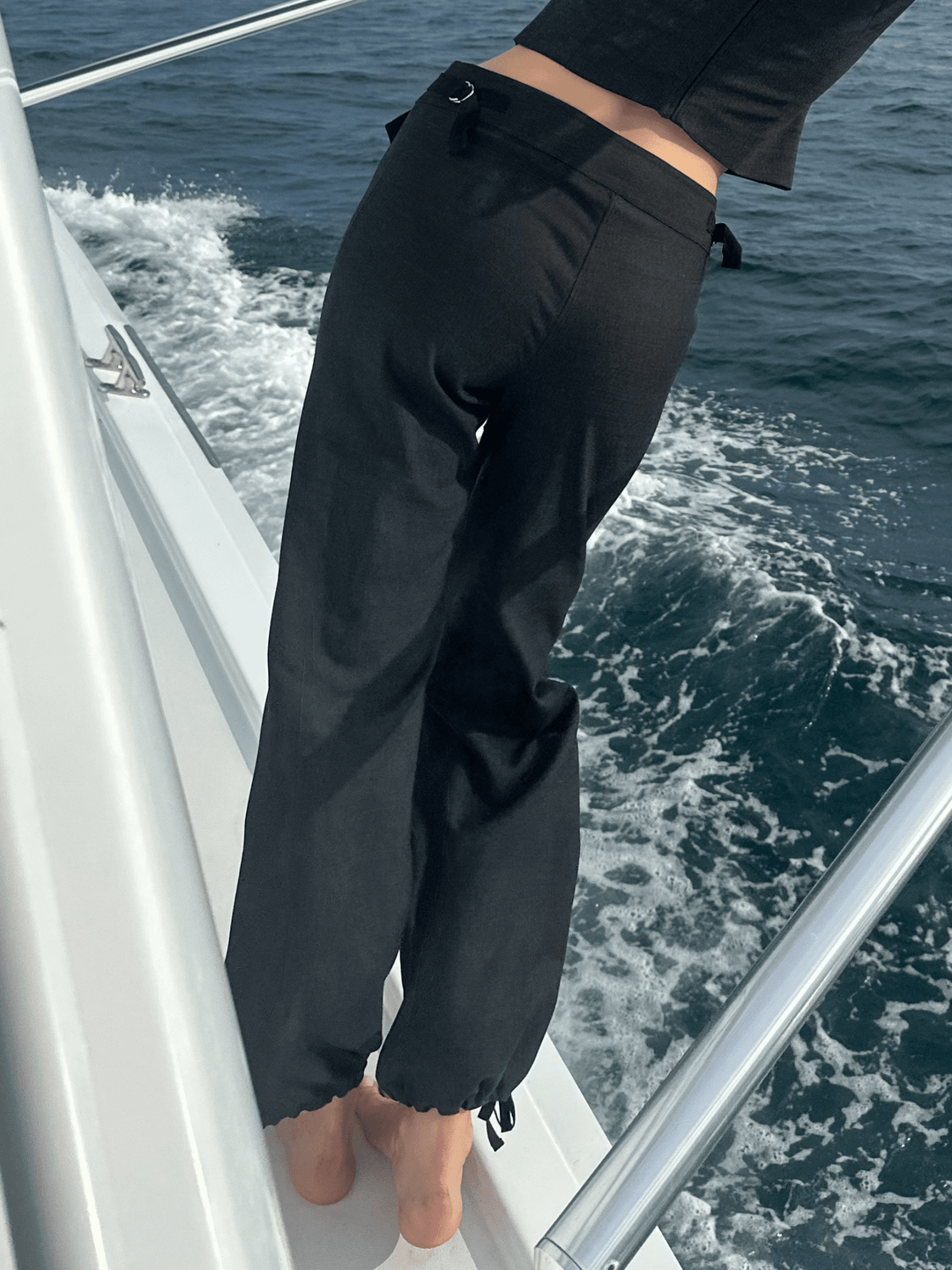 Model wearing black relaxed-fit pants with adjustable drawstrings at the waist, standing barefoot on the deck of a boat with the ocean in the background.