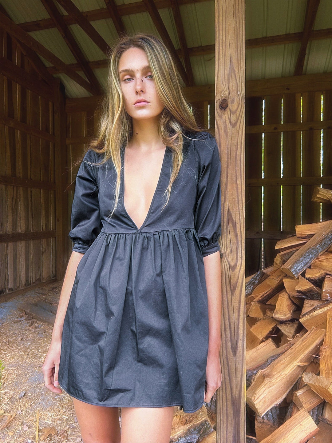 Model wearing a dark gray mini dress with a deep V-neckline and three-quarter length puff sleeves. The dress has a gathered waistline, creating a soft, flowing silhouette. The setting is a rustic wooden structure with stacked firewood in the background.