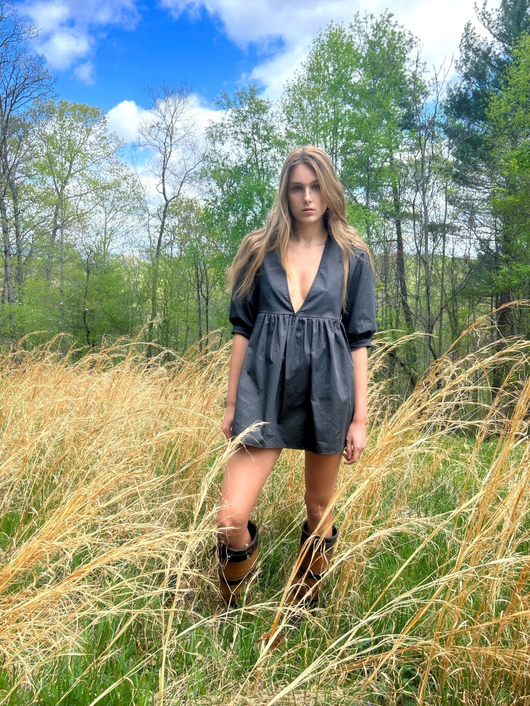 Model wearing a deep V-neck dark gray mini dress with puffed half sleeves, standing in a field of tall golden grass with a backdrop of green trees and a blue sky. Paired with knee-high brown and black boots.