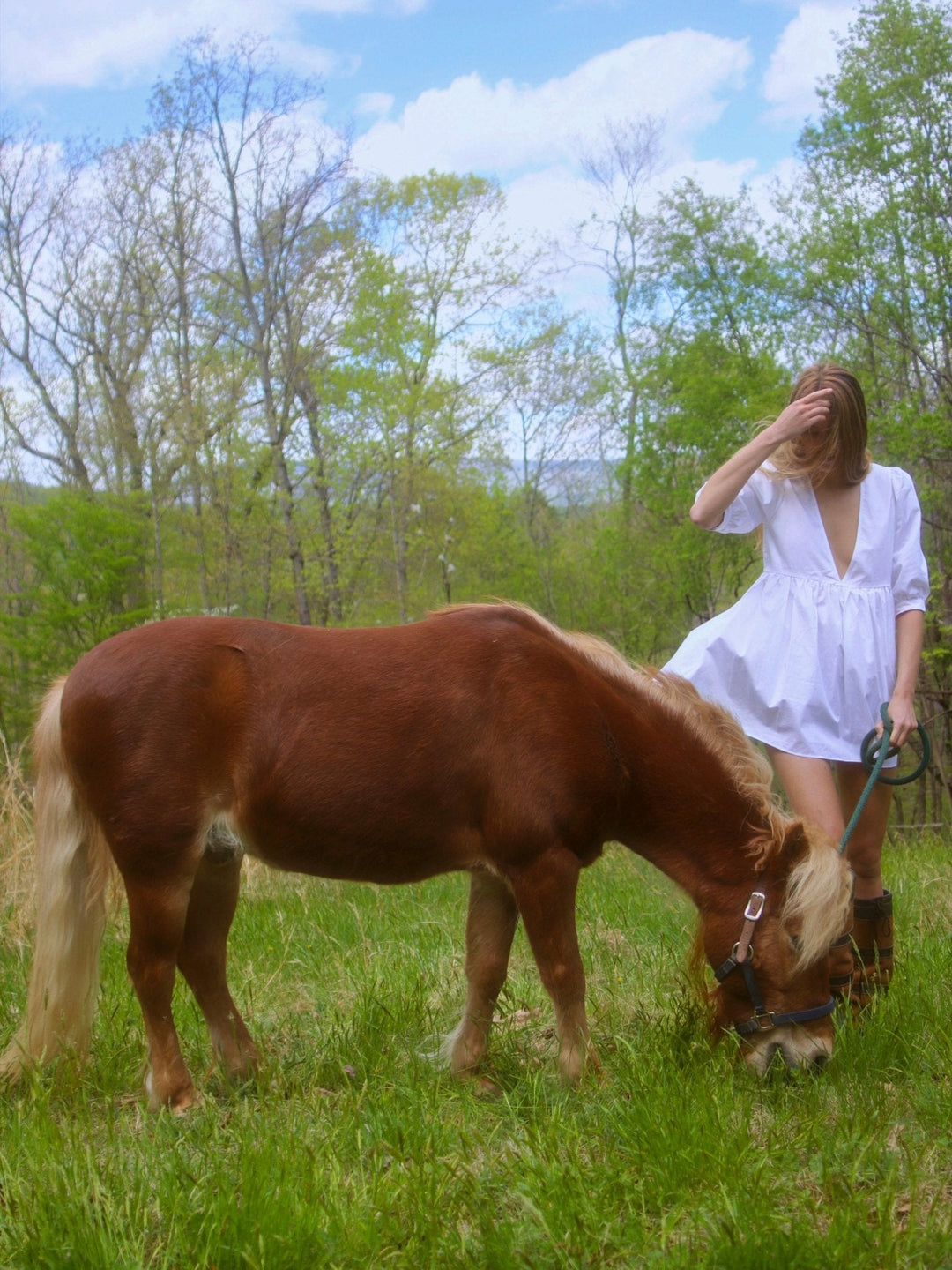 Model in a white dress stands in a grassy field holding a lead rope attached to a brown pony.