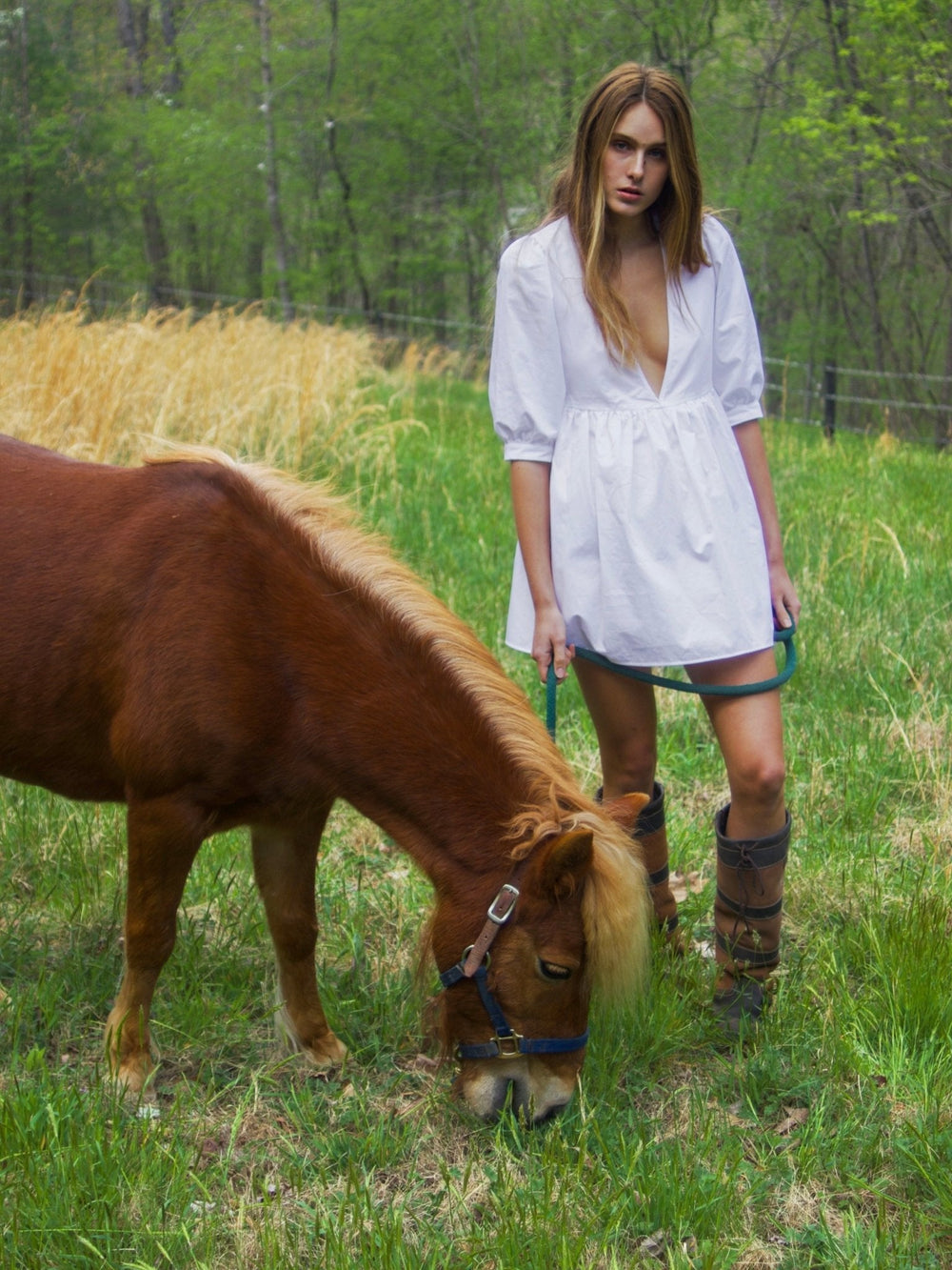 Model wearing a white dress with a deep V-neckline stands beside a brown pony. The background is filled with green trees, giving a serene, natural setting.