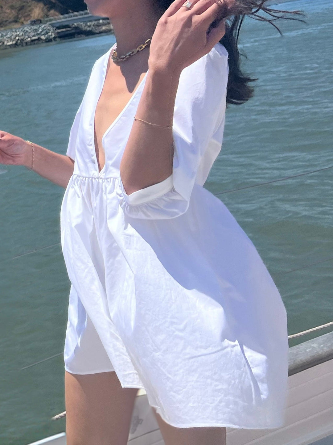 Model wearing a white dress with a deep V-neckline, standing by the water with wind blowing through her hair. 