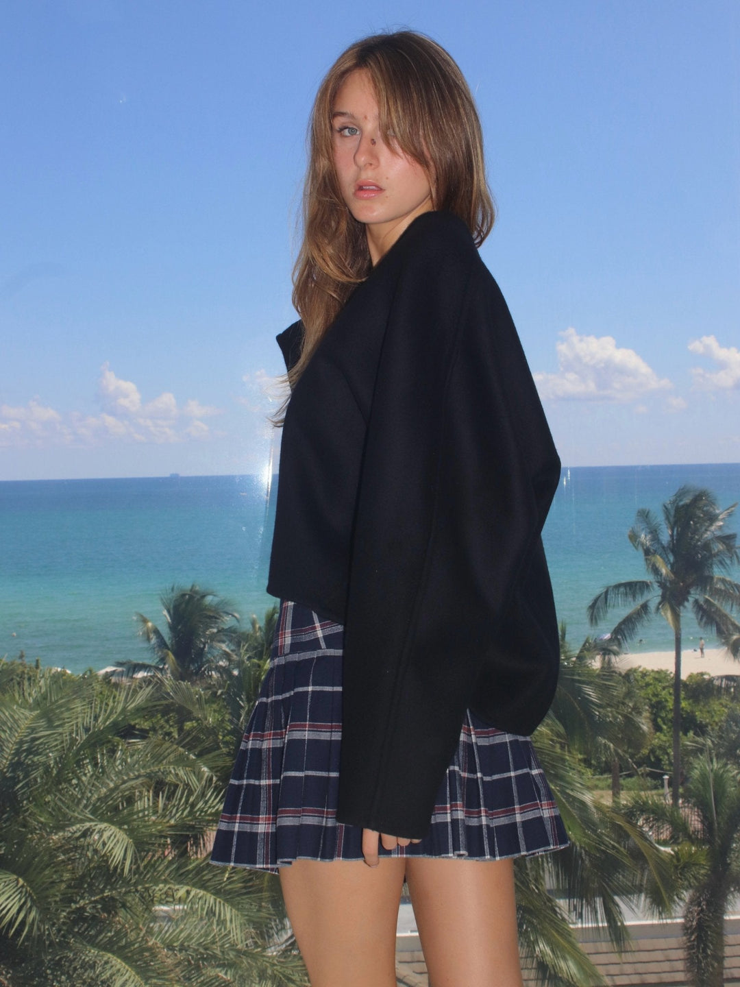 Model wearing a navy blue plaid pleated mini skirt and an oversized black sweater, with palm trees and the ocean in the background.