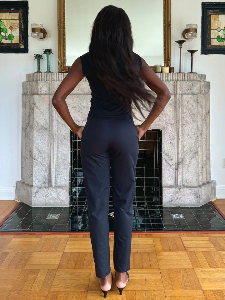 Back view of a model standing in front of a decorative fireplace, wearing a black sleeveless top, black pants, and heels.