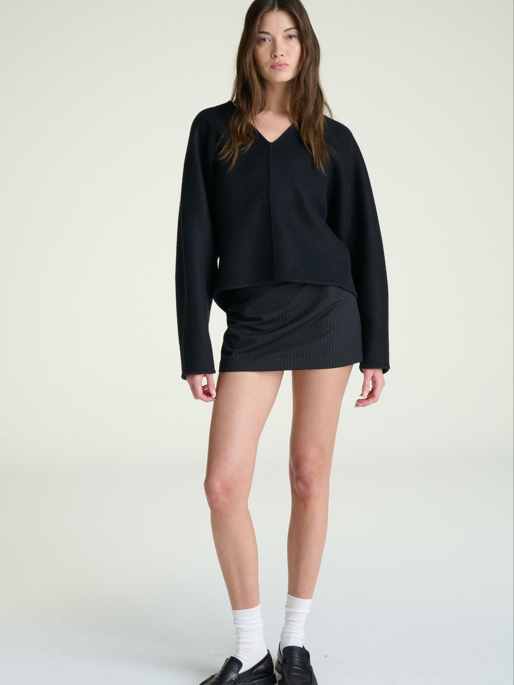 model wearing a black long-sleeve top and skirt with white socks and black shoes on a light background