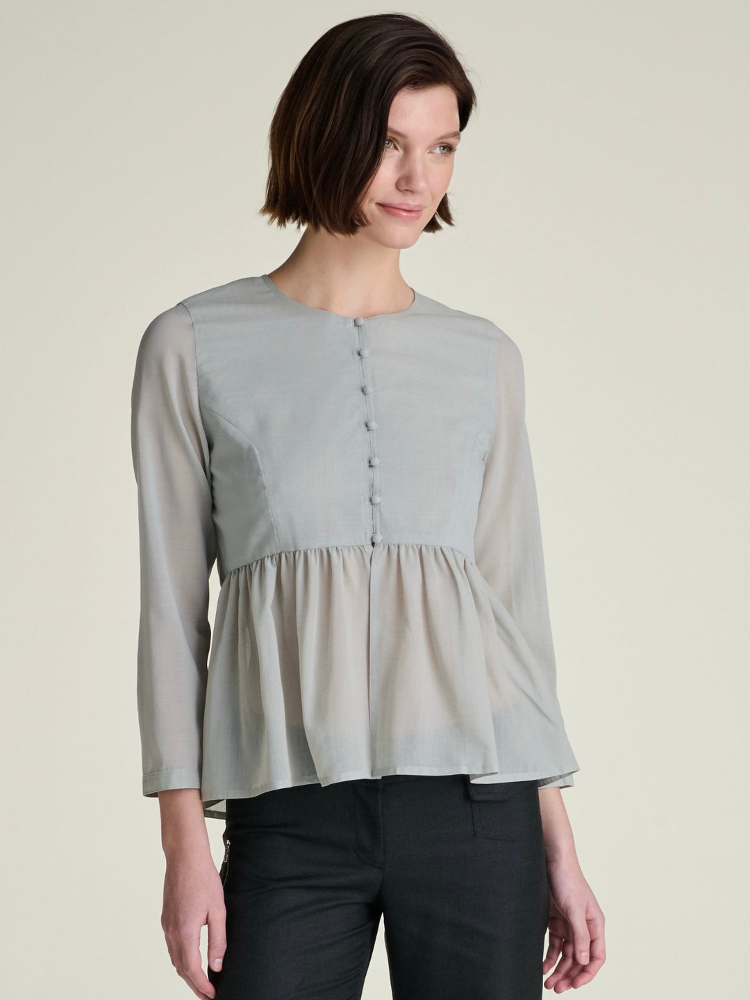 woman with short hair wearing a light grey-green blouse and black pants on a grey background