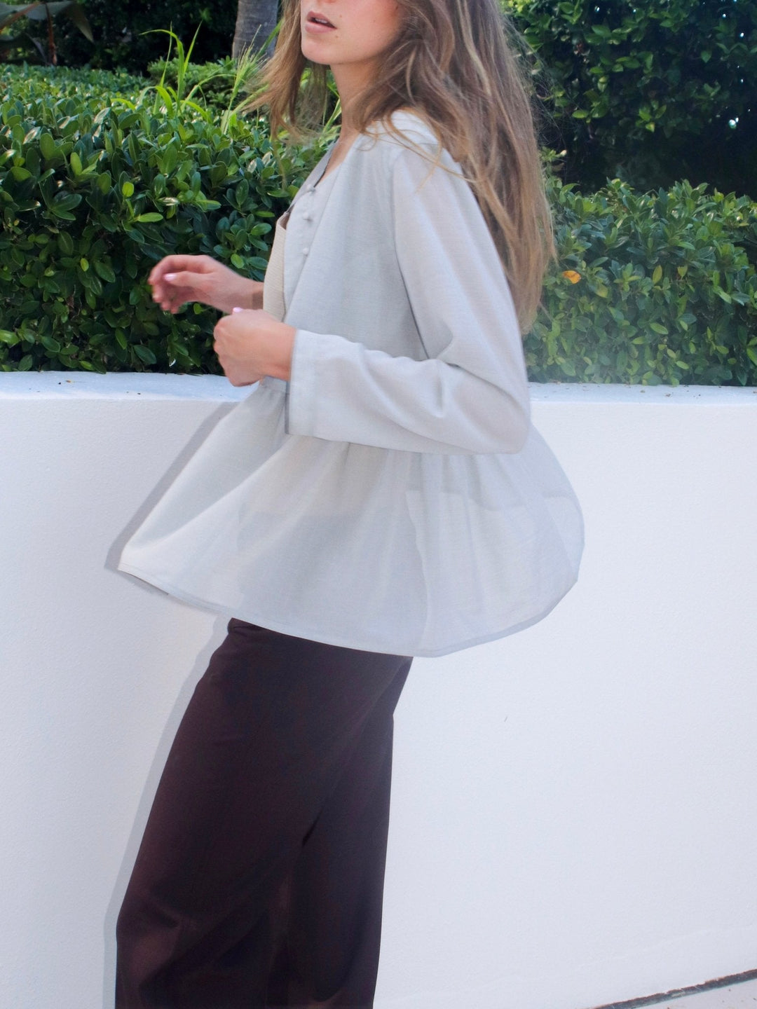 Model wearing a light grey, long-sleeved, flowing top with a flared hem. She pairs it with dark brown pants.