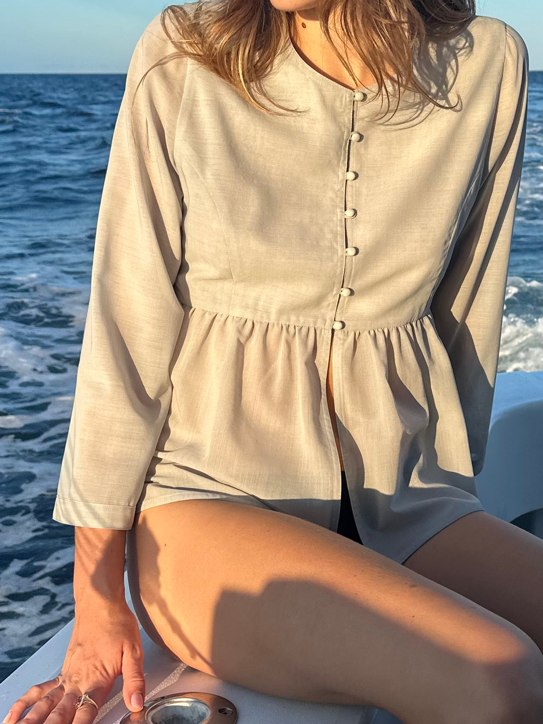 Close-up of a model wearing a light gray buttoned peplum blouse, sitting on a boat with the ocean in the background under natural sunlight.