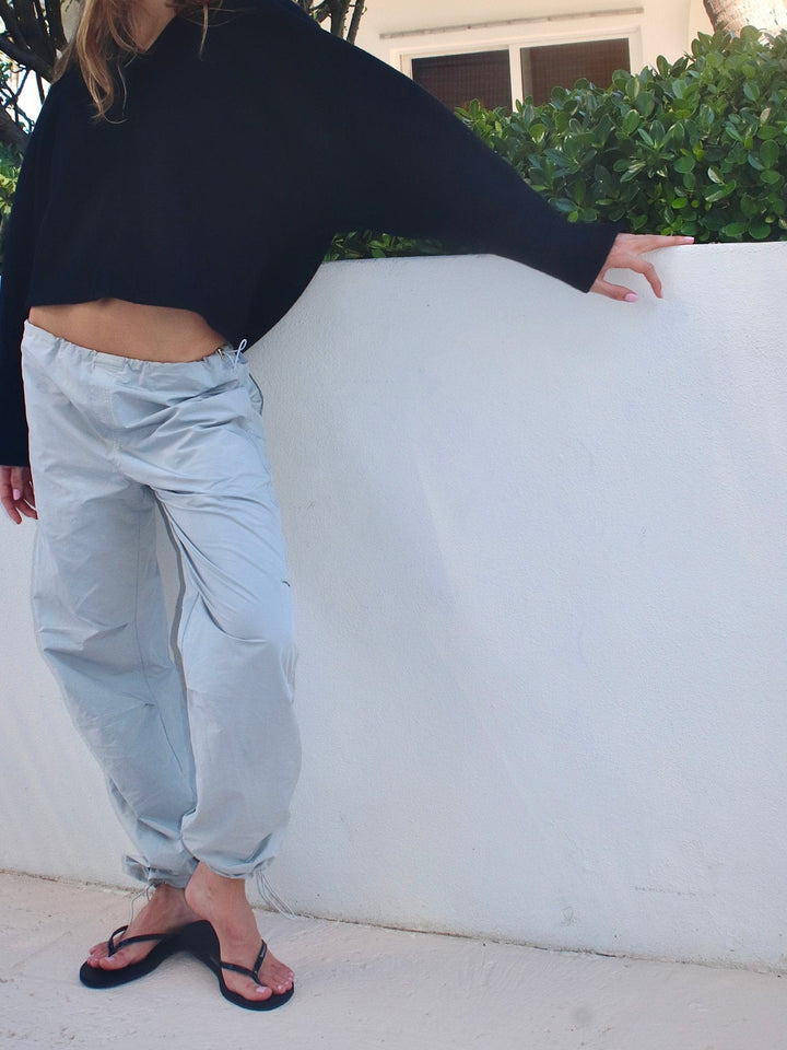 Model leaning against a white wall, dressed in light grey drawstring pants and a loose-fitting black top.