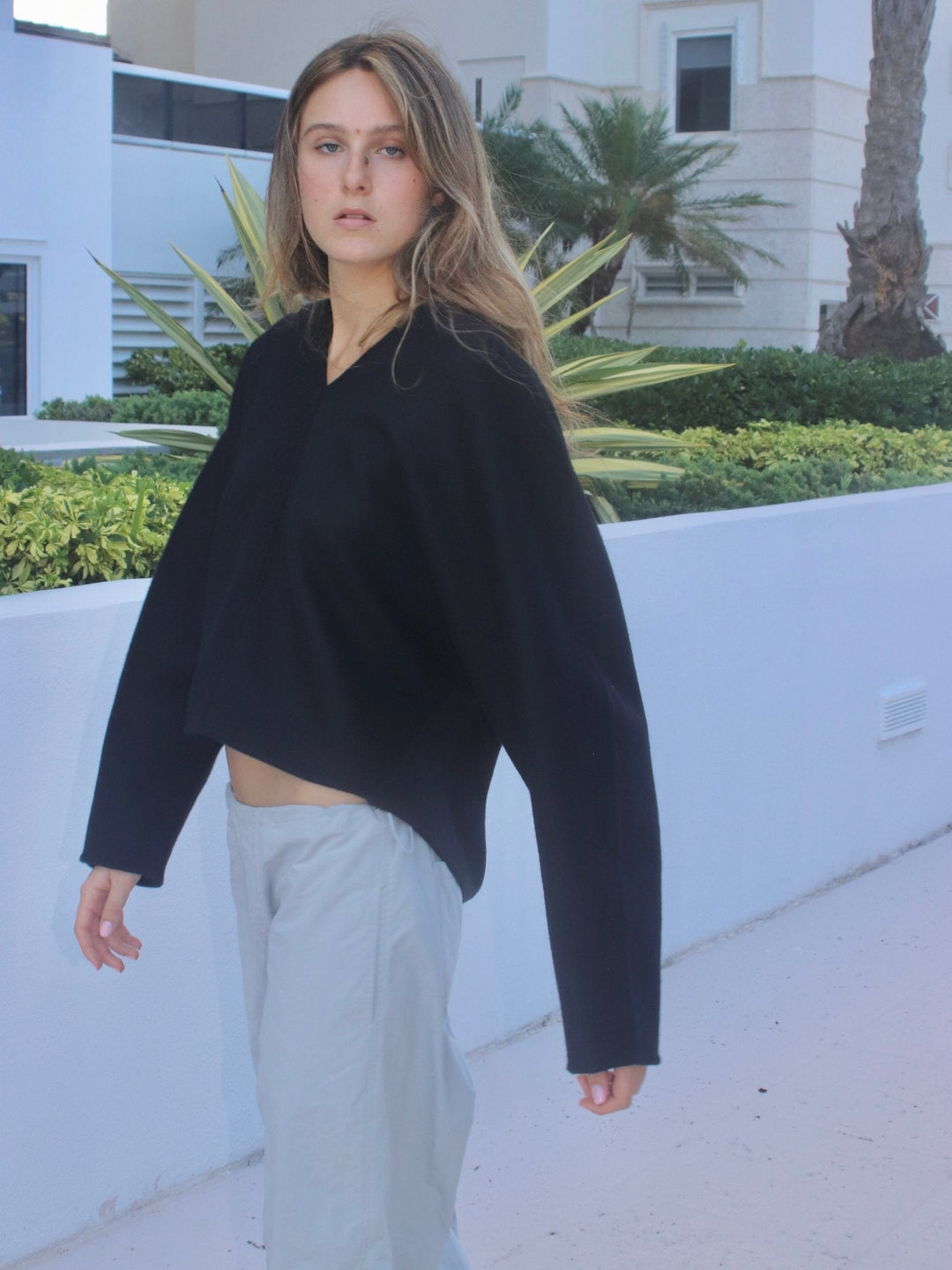 Model standing in a light outdoor setting, dressed in a black oversized sweater. They are also wearing light grey drawstring pants with a relaxed fit. In the background, there are green shrubs, palm trees, and a white building.