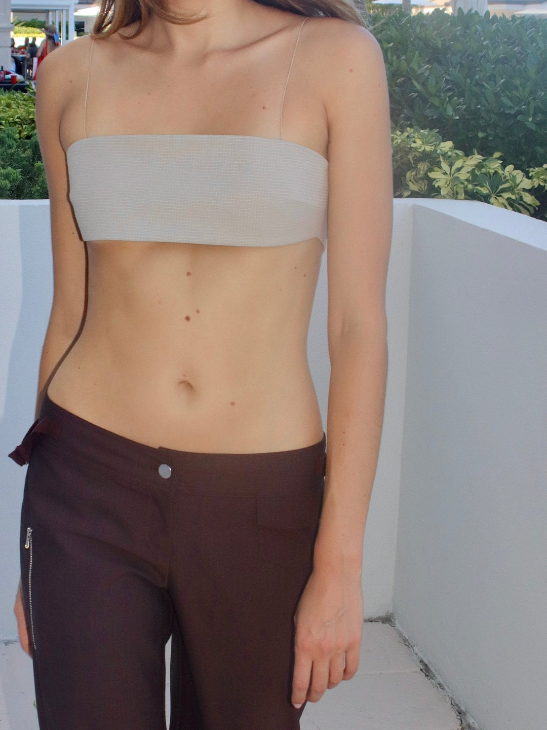 Model wearing a khaki bandeau top and brown pants, standing outdoors against a white wall with greenery in the background.