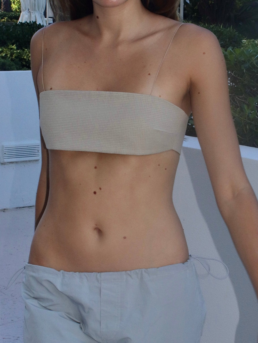 Model wearing a khaki bandeau top and light gray pants, standing outdoors with a white wall and greenery in the background.