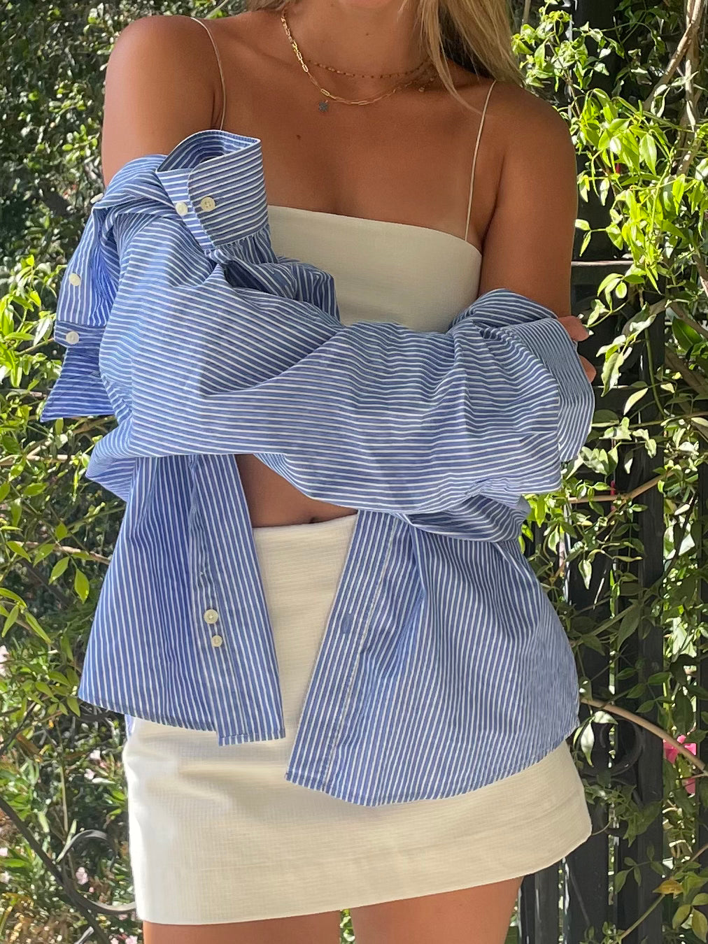 Model wearing a blue striped oversized shirt over a white bandeau top and mini skirt, posing outdoors with greenery in the background.