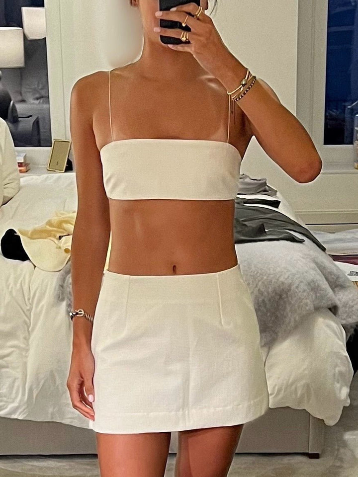 Model wearing a white, strapless crop top with thin spaghetti straps, paired with a matching white high-waisted mini skirt. 