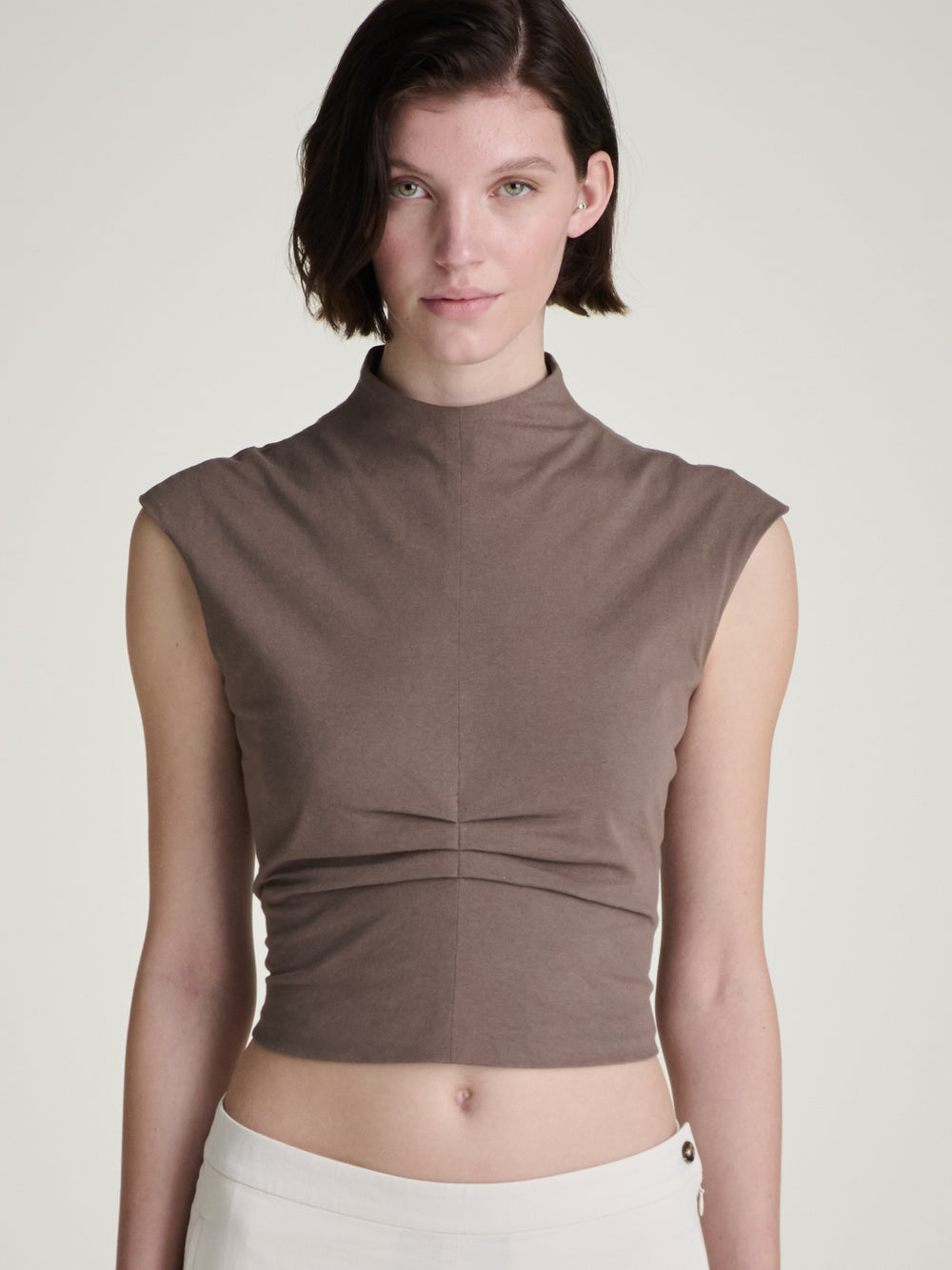 A woman wearing a sleeveless brown mockneck top on a light grey background