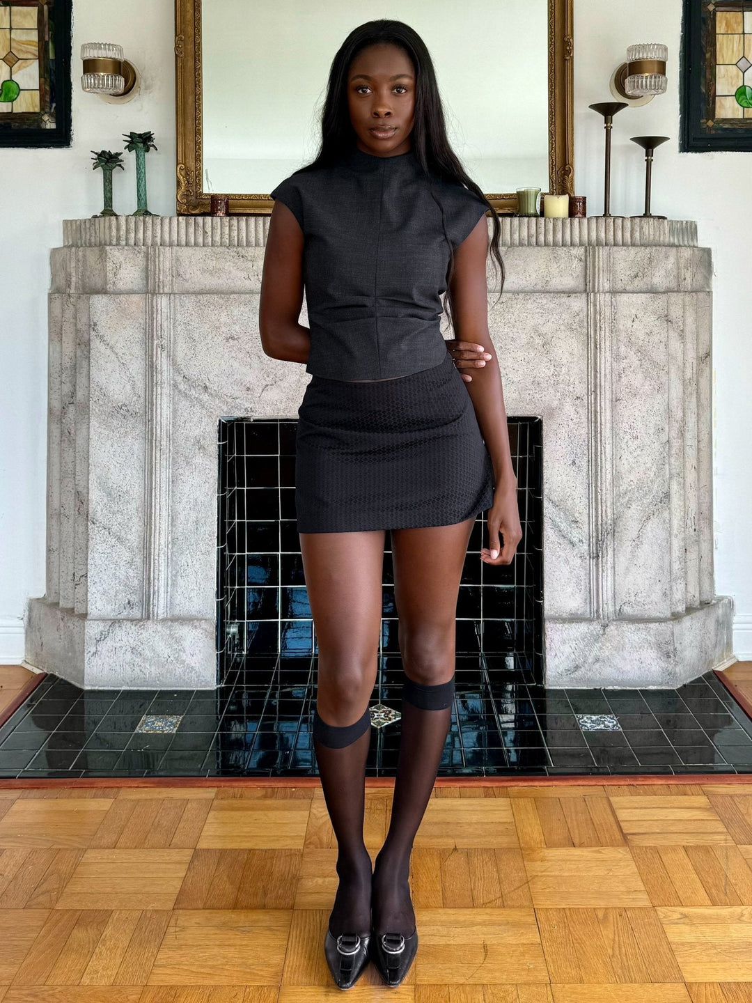 Model wearing a fitted charcoal sleeveless top, a black textured mini skirt, knee-high sheer socks, and black loafers, standing indoors in front of a decorative fireplace.