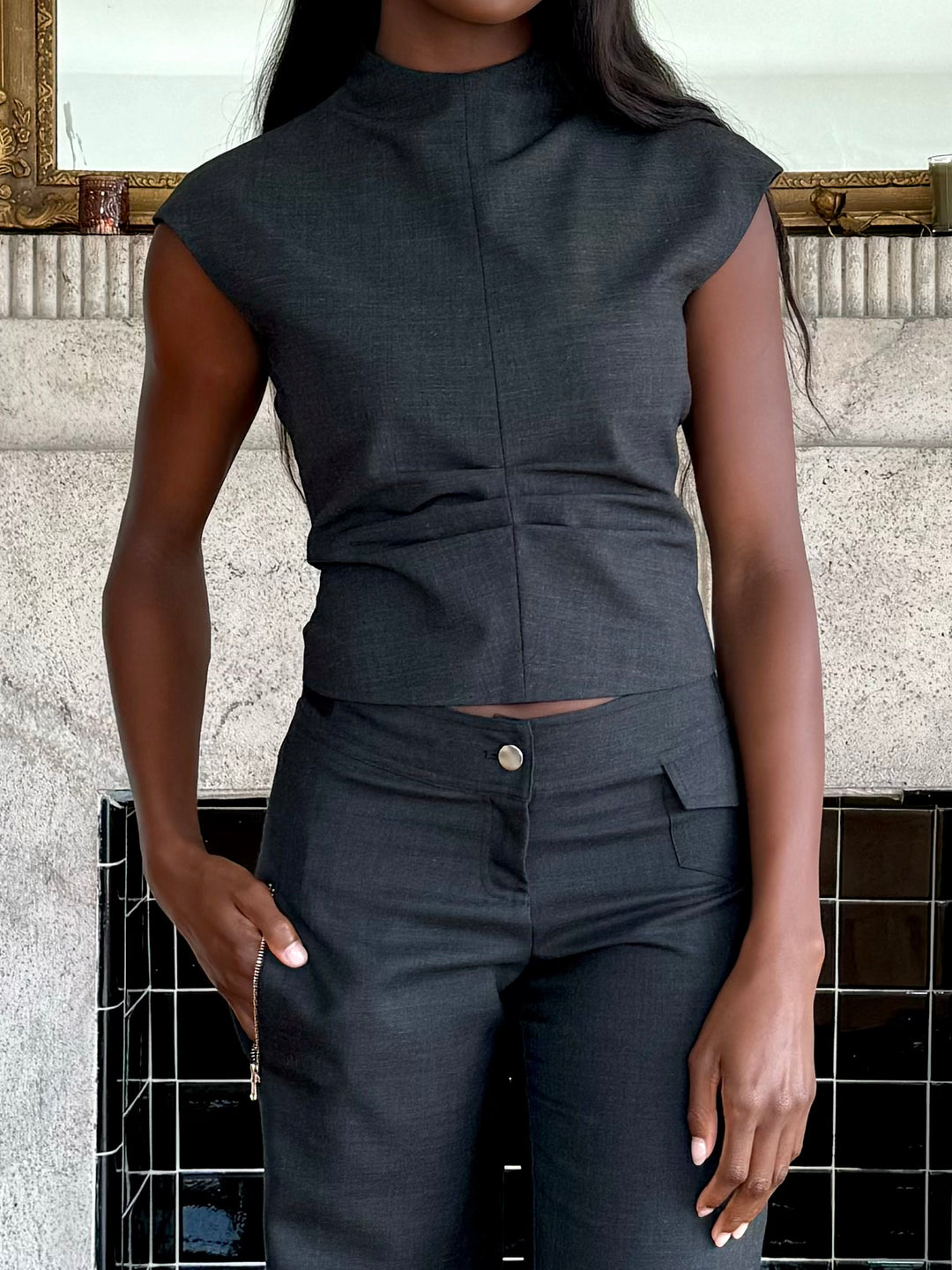 Model wearing a fitted charcoal sleeveless top and matching charcoal pants with a button closure, standing indoors in front of a decorative fireplace.