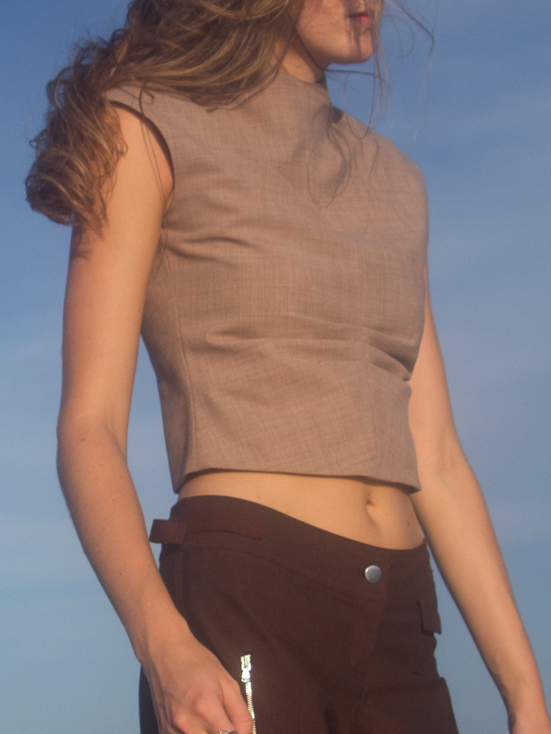 Model wearing a beige sleeveless turtleneck top paired with brown pants, standing outdoors with a clear blue sky in the background.