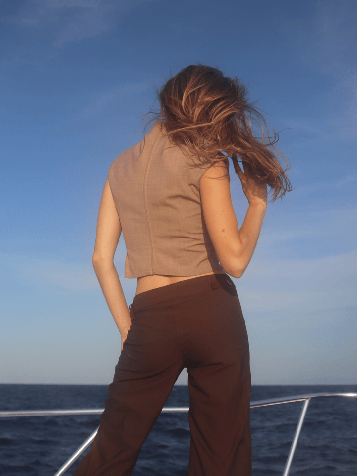 Back view of a woman on a boat wearing a beige sleeveless top and brown pants. She stands against a backdrop of ocean and sky, with her hair blowing in the wind.