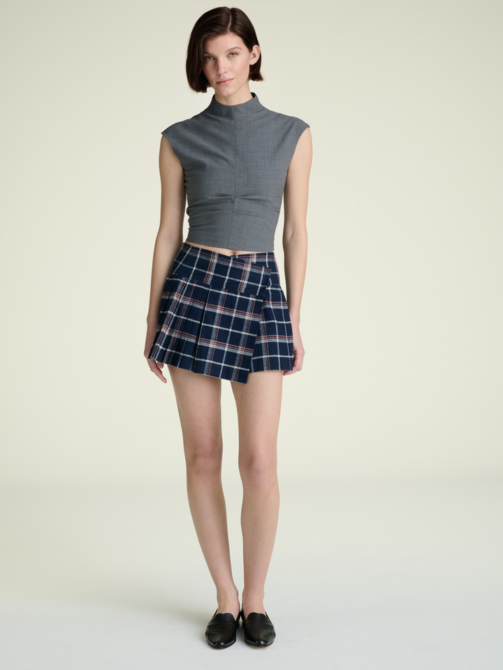 full body woman with short hair wearing a grey top with a mockneck collar and cap sleeves and wearing a plaid skirt and black shoes