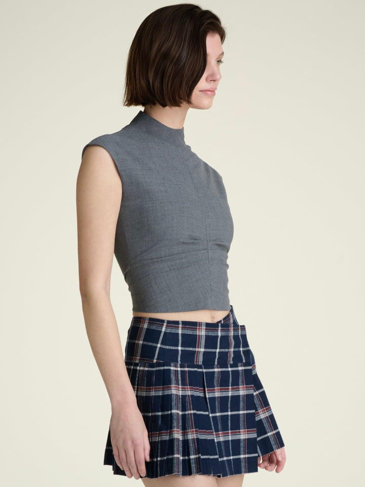 woman in three quarter profile wearing a grey mockneck and cap sleeve wool top and plaid skirt on a grey background
