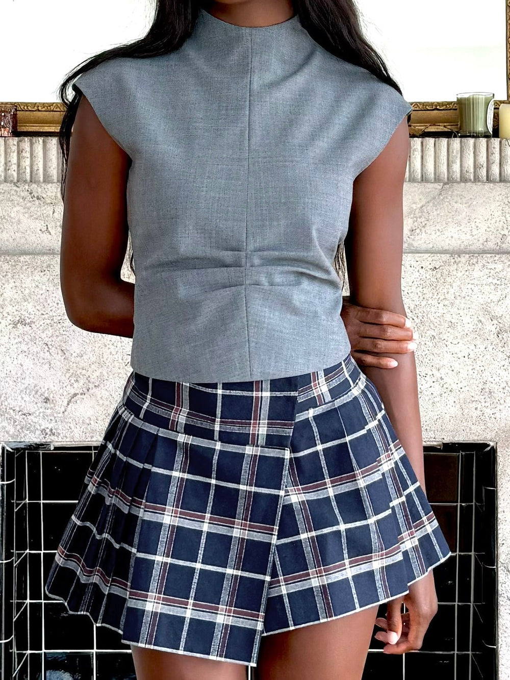 Model wearing a grey sleeveless top with a navy plaid pleated skirt. She stands in front of a stone fireplace.