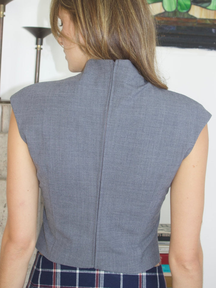 Back view of a gray sleeveless high neck top, showing a seam down the center. The model pairs it with a navy plaid pleated skirt.