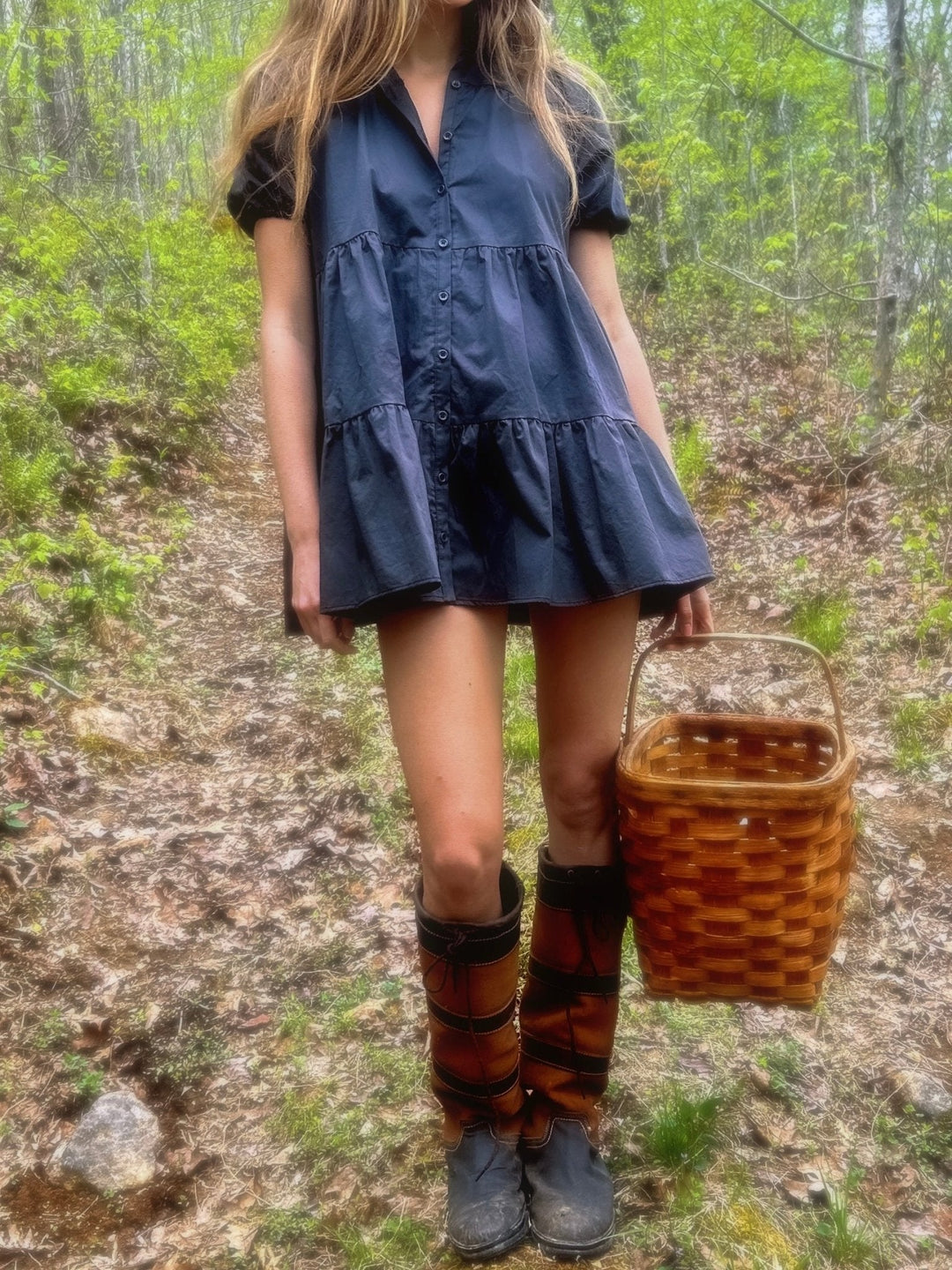 Model in a forest, wearing a slate short dress with button-down front and ruffled hem. She pairs the dress with knee-high boots and holds a wicker basket.