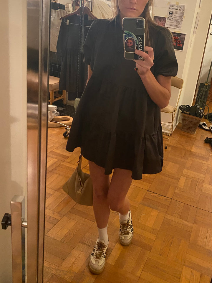 Mirror selfie of a person wearing a black tiered dress with puff sleeves, white socks, and beige Adidas sneakers. 