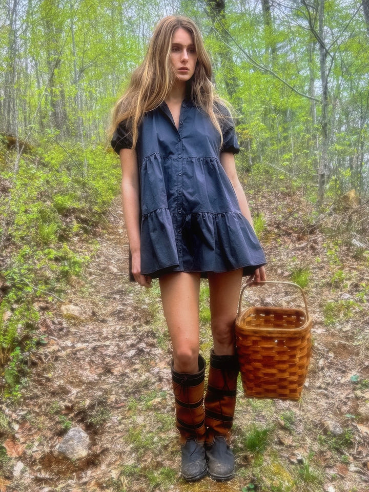 Model in a forest, wearing a slate short dress with button-down front and ruffled hem. She pairs the dress with knee-high boots and holds a wicker basket.