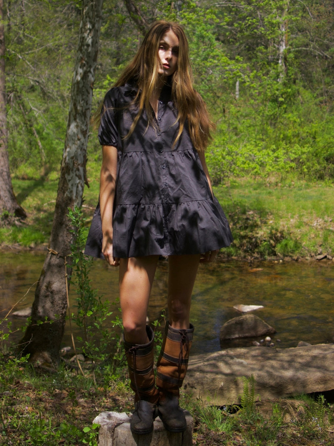 Model standing by a stream wearing a slate ruffled mini dress with a button-down front. She compliments her outfit with brown knee-high boots.