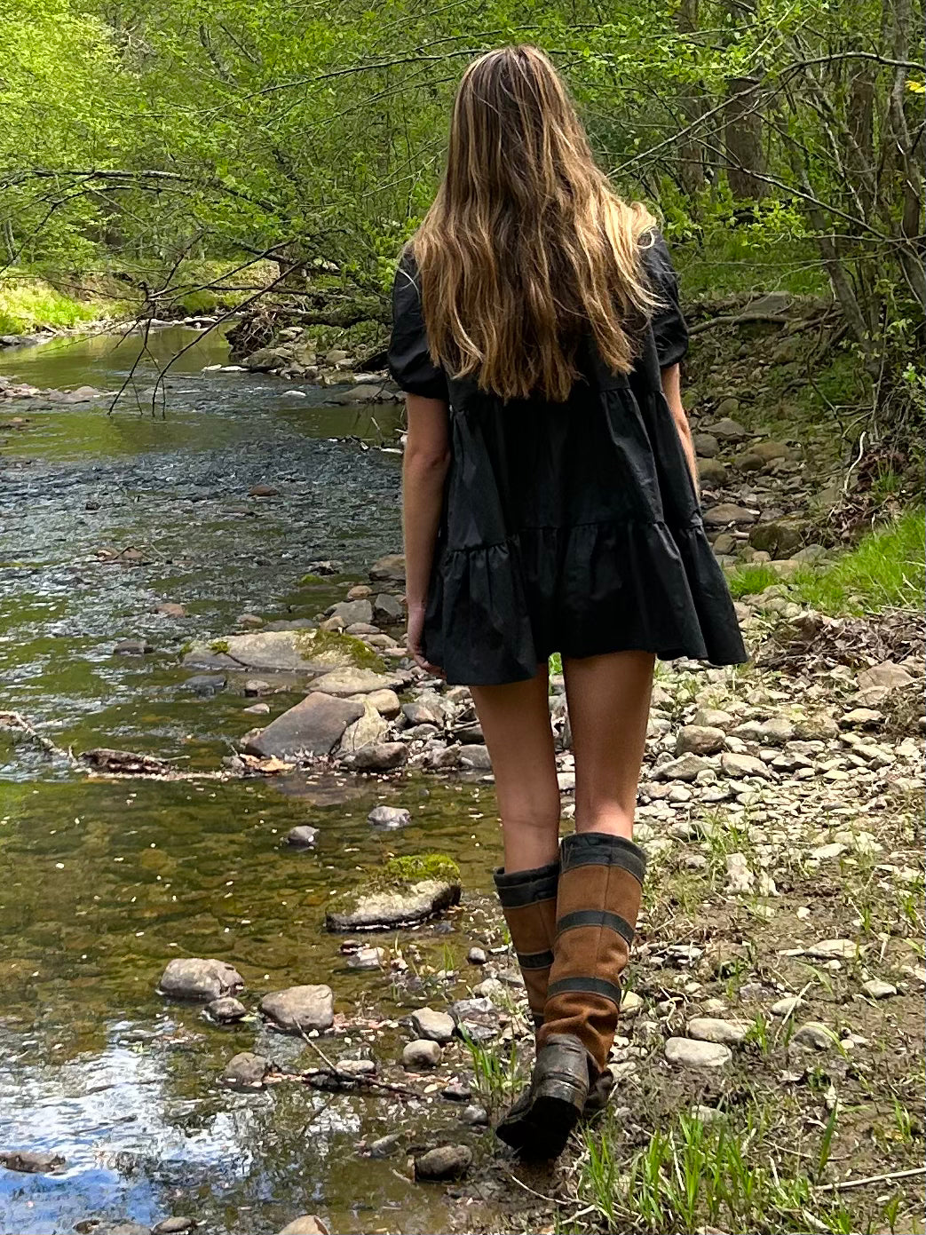 Back view of a model wearing a black tiered mini-dress with knee-high leather boots, walking along a rocky stream.