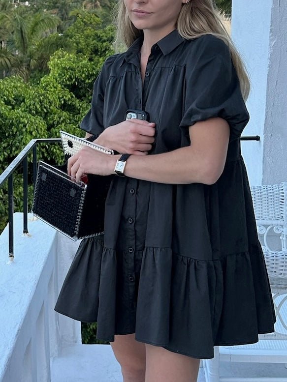 Model wearing a slate button-down dress with short puff sleeves, holding a textured black clutch and a phone. She stands on a balcony with a white railing and greenery in the background.