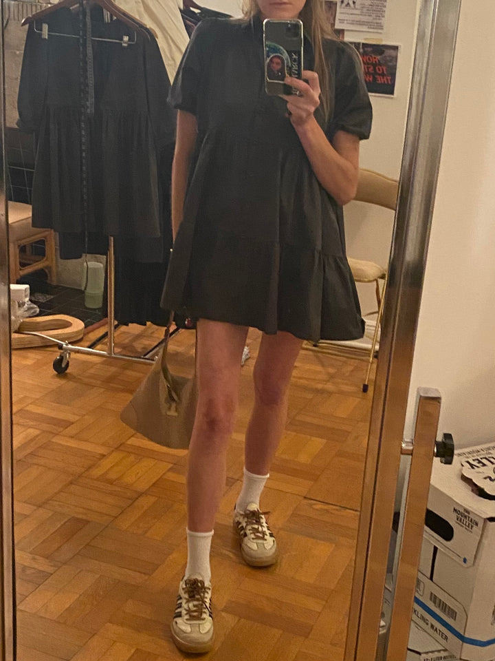 Mirror selfie of a person wearing a black tiered dress with puff sleeves, white socks, and beige Adidas sneakers. 