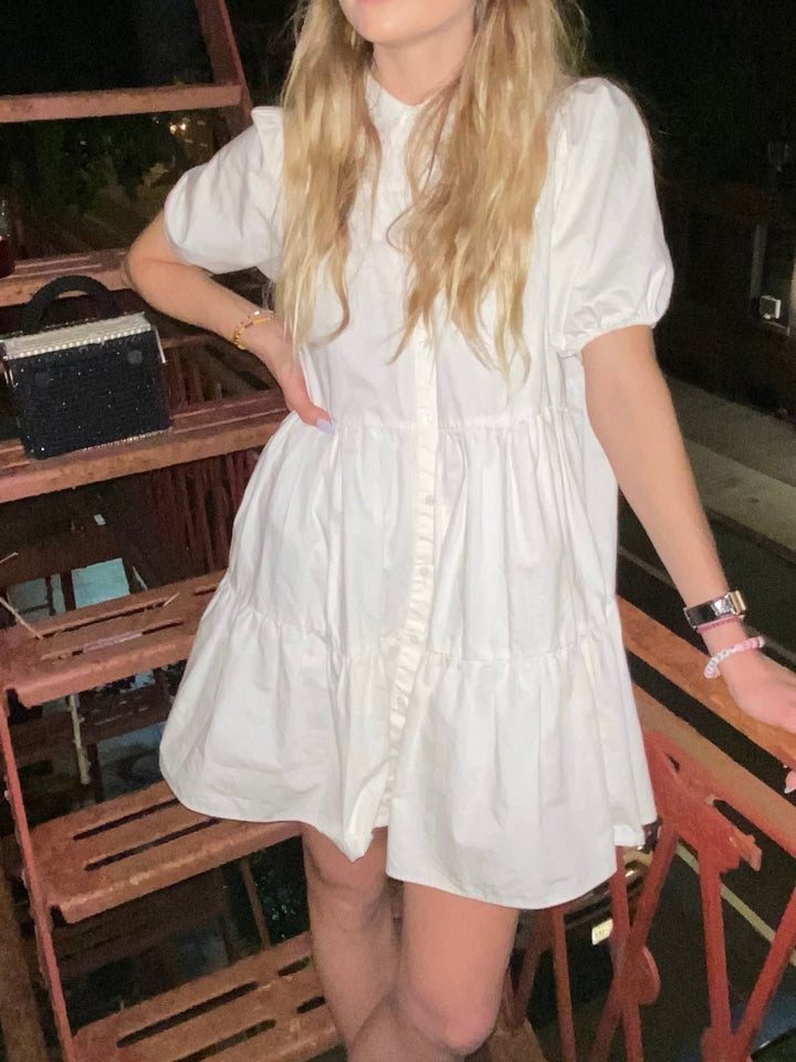 Model wearing a white mini dress with short puff sleeves and tiered ruffles. She poses on a wooden deck at night, accessorized with a sparkly black handbag and stylish wrist watch.