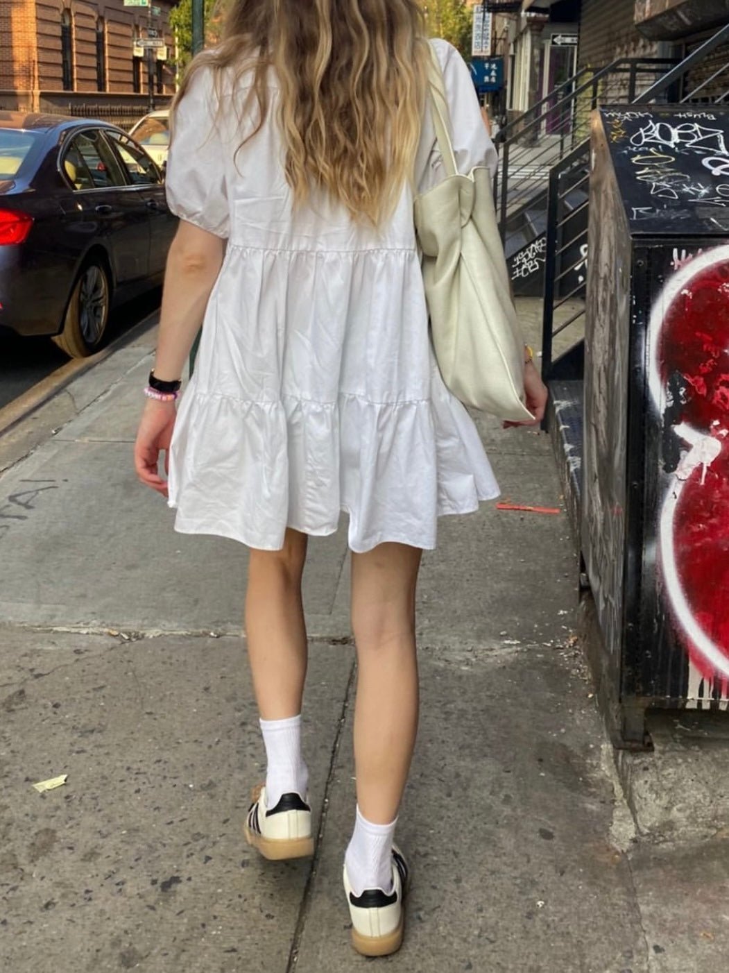 Rear view of a woman walking down a city street, wearing a short white dress with tiered ruffles and puff sleeves. She pairs the dress with white socks and white shoes, carrying a light beige shoulder bag.