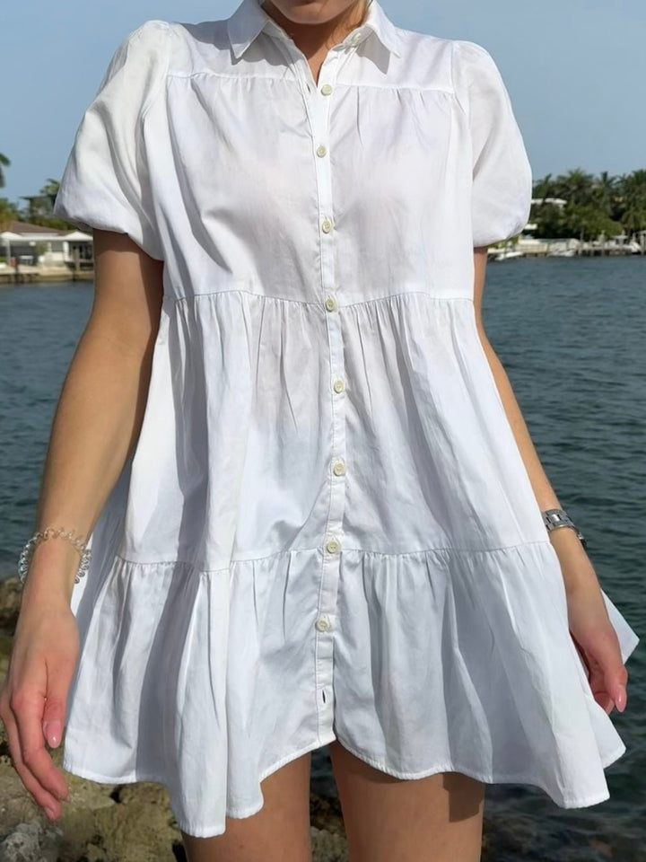 Model wearing a white dress with short sleeves, a collar, and buttons down the front. She's standing by the water on a sunny day.