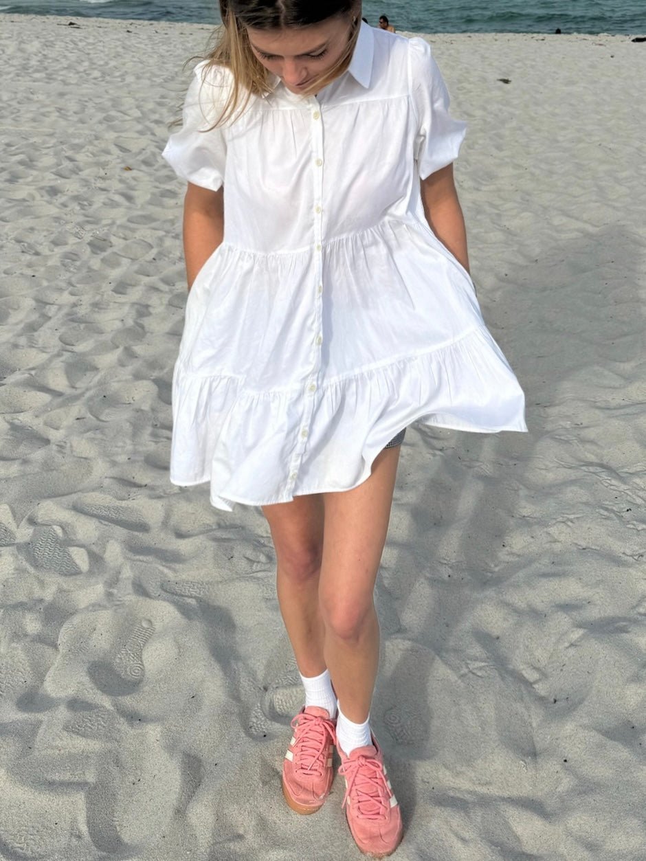 Model wearing a white button-down dress with short sleeves, standing on a sandy beach. She pairs the dress with pink sneakers and white socks.