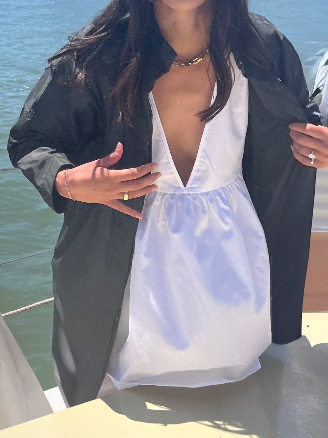 Model standing on a boat, styling a deep V-neck white dress under an open black jacket with the ocean in the background.